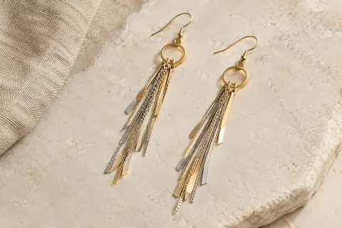 Paire de boucles pendantes à longues pampilles dans des tons doré et argenté, posée sur une surface claire.
