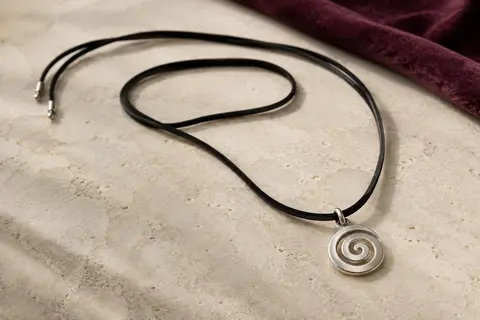 Sautoir à pendentif en spirale argentée et cordon noir en suédine posé sur une surface claire.