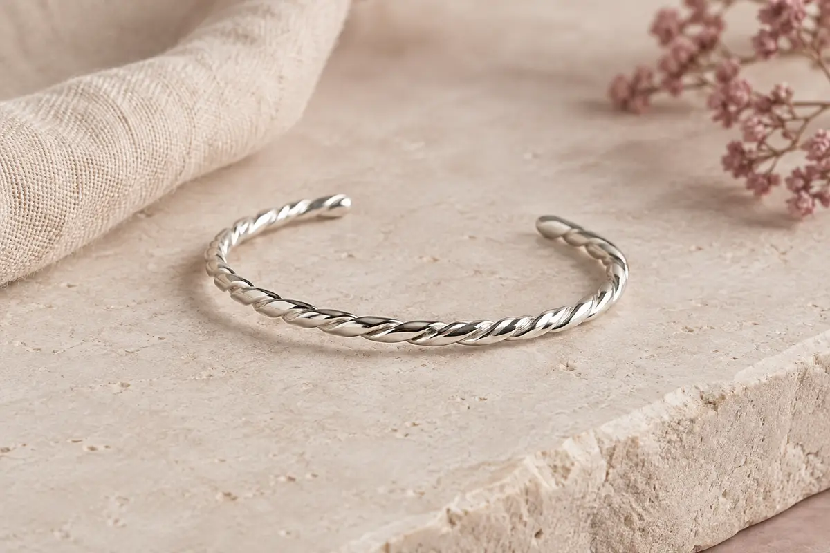 Bracelet jonc ouvert torsadé au fini argenté posé seul sur une surface minérale claire.
