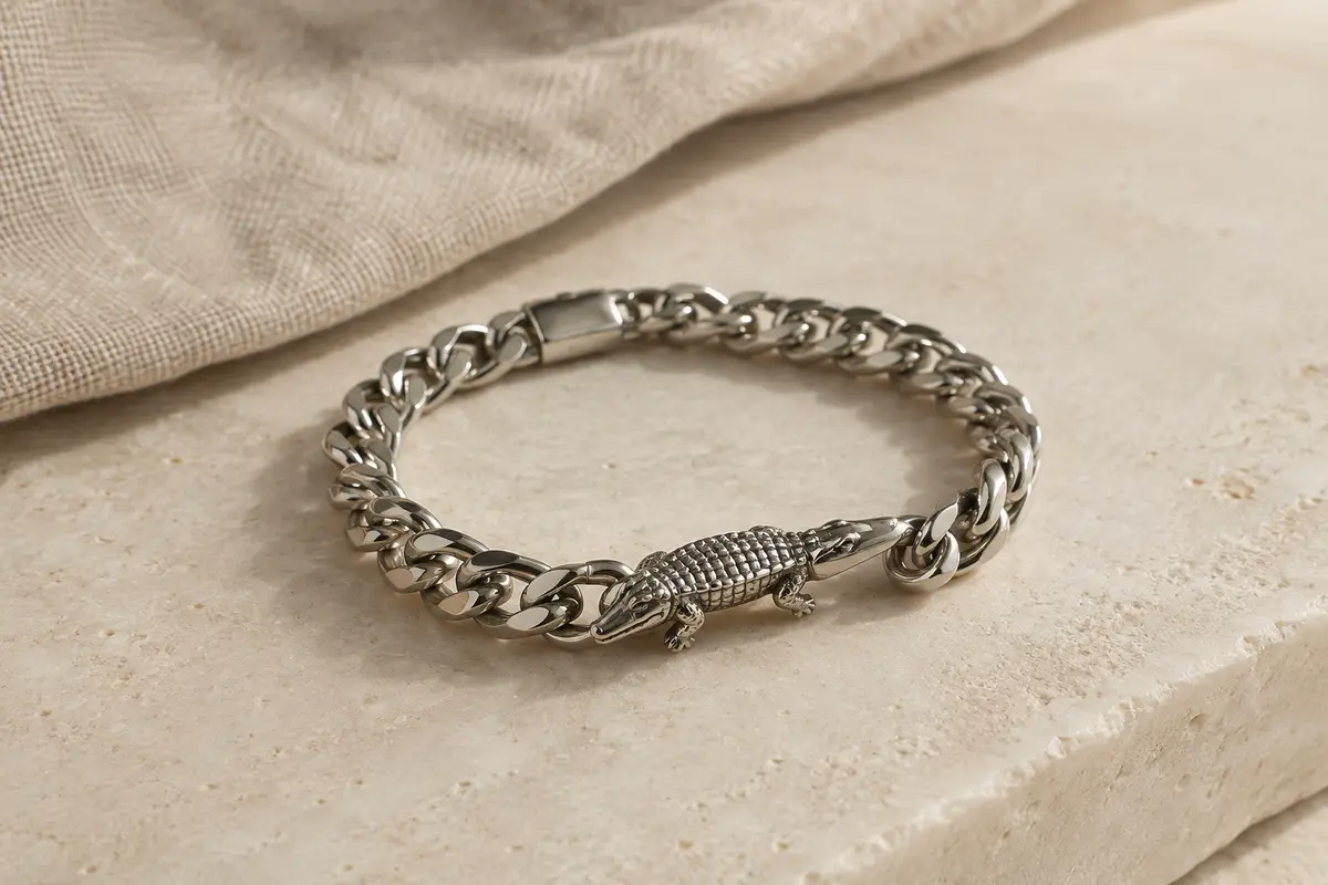 Bracelet chaîne argenté à gros maillons avec un petit détail crocodile, posé sur une surface ivoire.