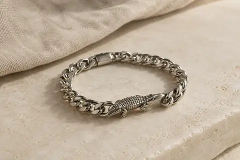 Bracelet chaîne argenté à gros maillons avec un petit détail crocodile, posé sur une surface ivoire.