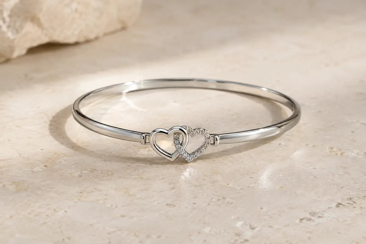 Bracelet jonc à finition rhodiée avec deux cœurs blancs reliés, posé sur une surface minérale claire.