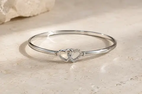 Bracelet jonc à finition rhodiée avec deux cœurs blancs reliés, posé sur une surface minérale claire.