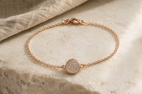 Bracelet à chaîne au ton or rosé avec un disque central de cristaux pavés, posé sur une surface minérale claire.