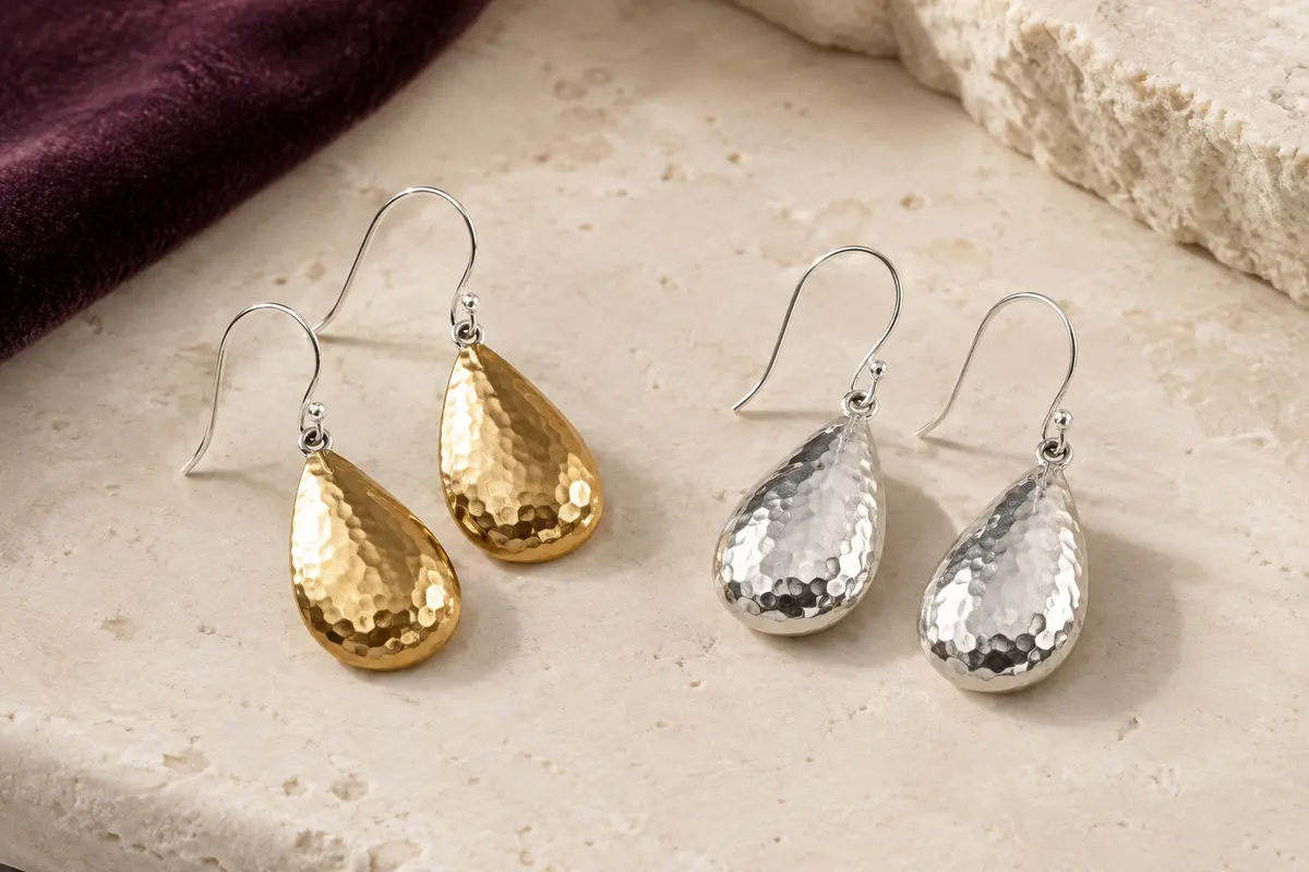 Boucles d’oreilles pendantes en goutte martelée, présentées en finitions dorée et argentée sur une surface ivoire
