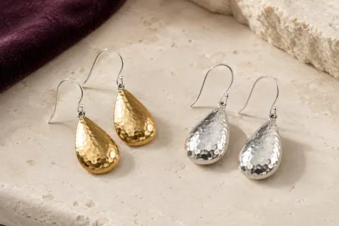 Boucles d’oreilles pendantes en goutte martelée, présentées en finitions dorée et argentée sur une surface ivoire