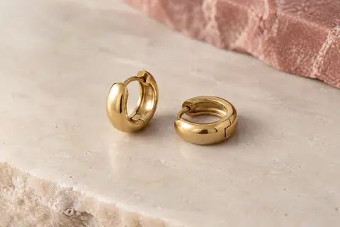 Petites créoles huggie dorées en cuivre, posées seules sur une surface minérale claire