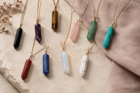 Douze colliers à pendentifs hexagonaux colorés disposés sur une surface minérale claire et du lin