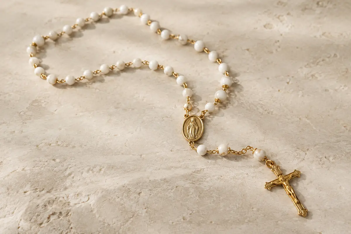 Collier chapelet doré à perles blanches, avec médaille mariale et crucifix, posé sur une surface claire.