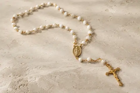 Collier chapelet doré à perles blanches, avec médaille mariale et crucifix, posé sur une surface claire.