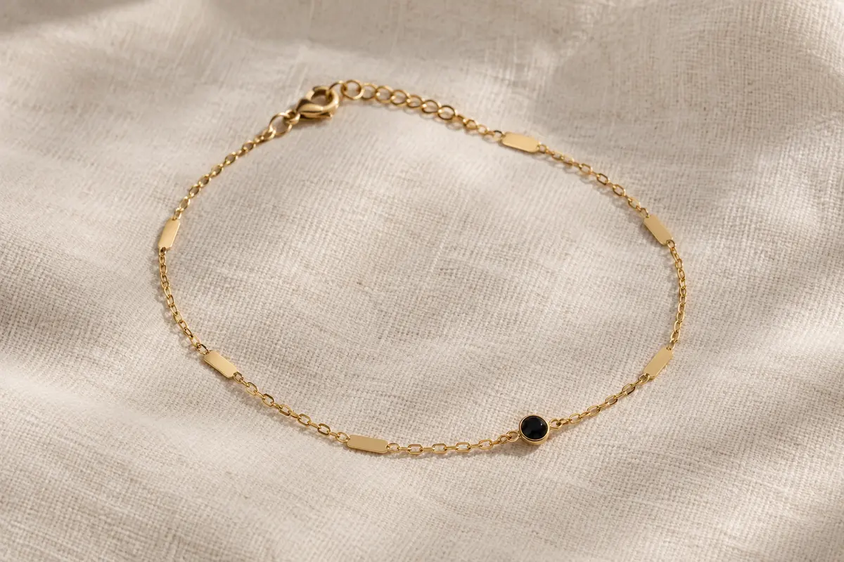 Bracelet de cheville doré à chaîne câble et petites plaques, posé seul sur du lin ivoire