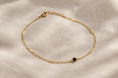 Bracelet de cheville doré à chaîne câble et petites plaques, posé seul sur du lin ivoire
