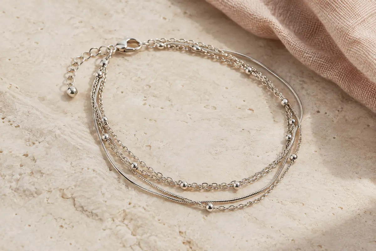 Bracelet de cheville multi-rangs à chaîne serpent posé sur une pierre claire près d’un tissu en lin naturel.