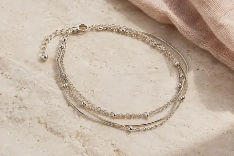 Bracelet de cheville multi-rangs à chaîne serpent posé sur une pierre claire près d’un tissu en lin naturel.