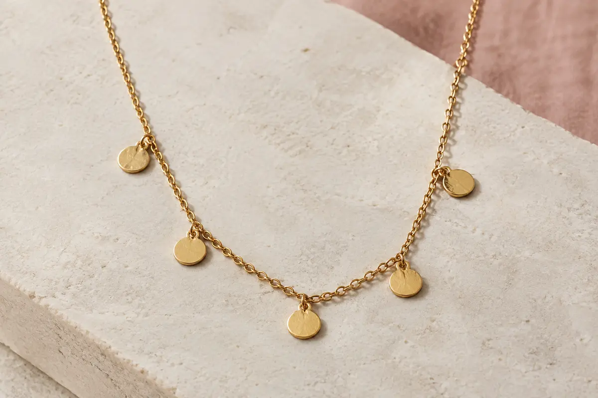 Collier doré à chaîne forçat et cinq petites pièces rondes, posé sur une surface minérale claire.