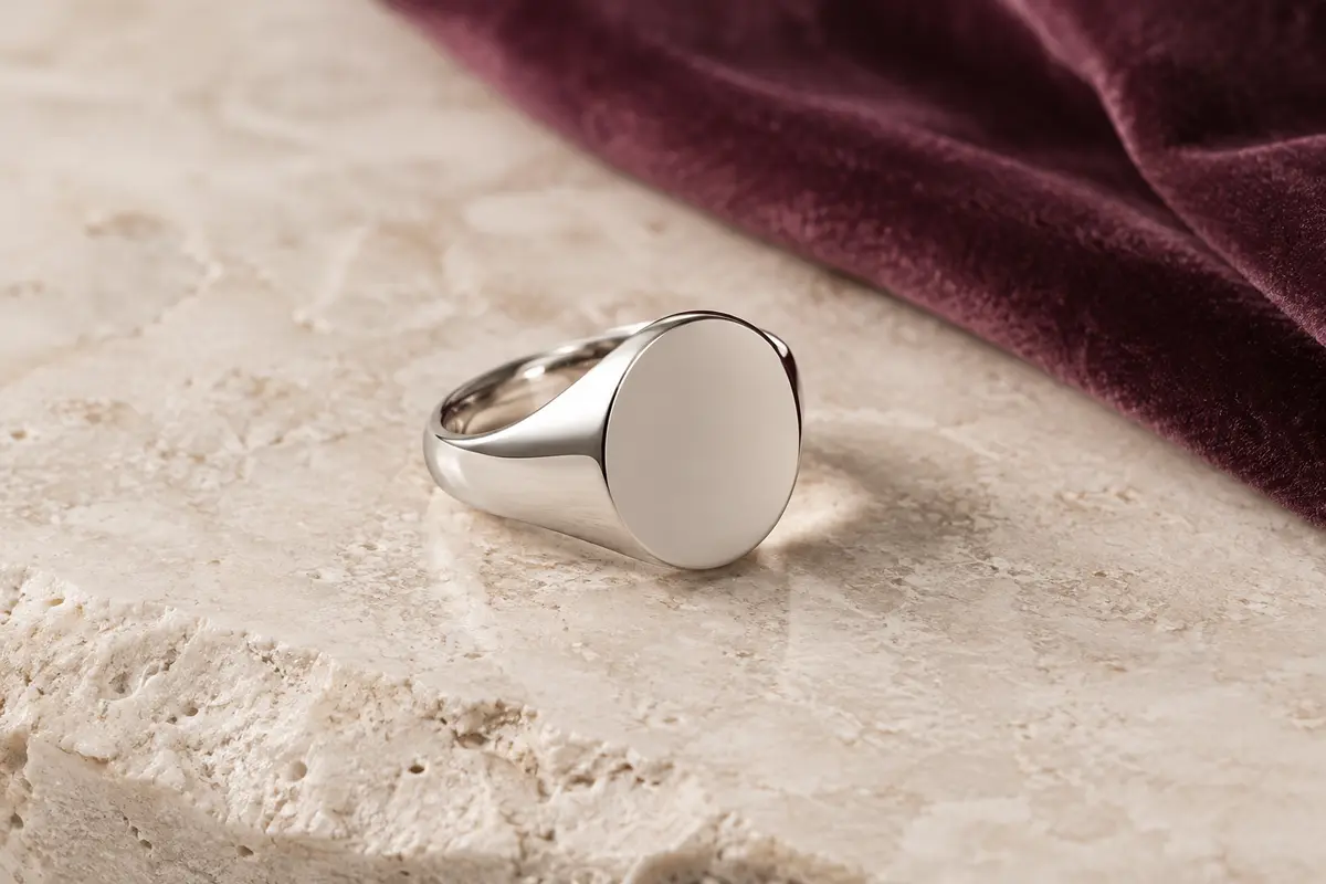 Chevalière ronde en acier inoxydable à la face polie, présentée sur une surface minérale claire avec du velours prune.