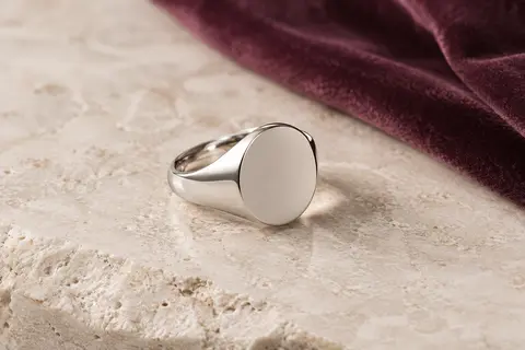 Chevalière ronde en acier inoxydable à la face polie, présentée sur une surface minérale claire avec du velours prune.