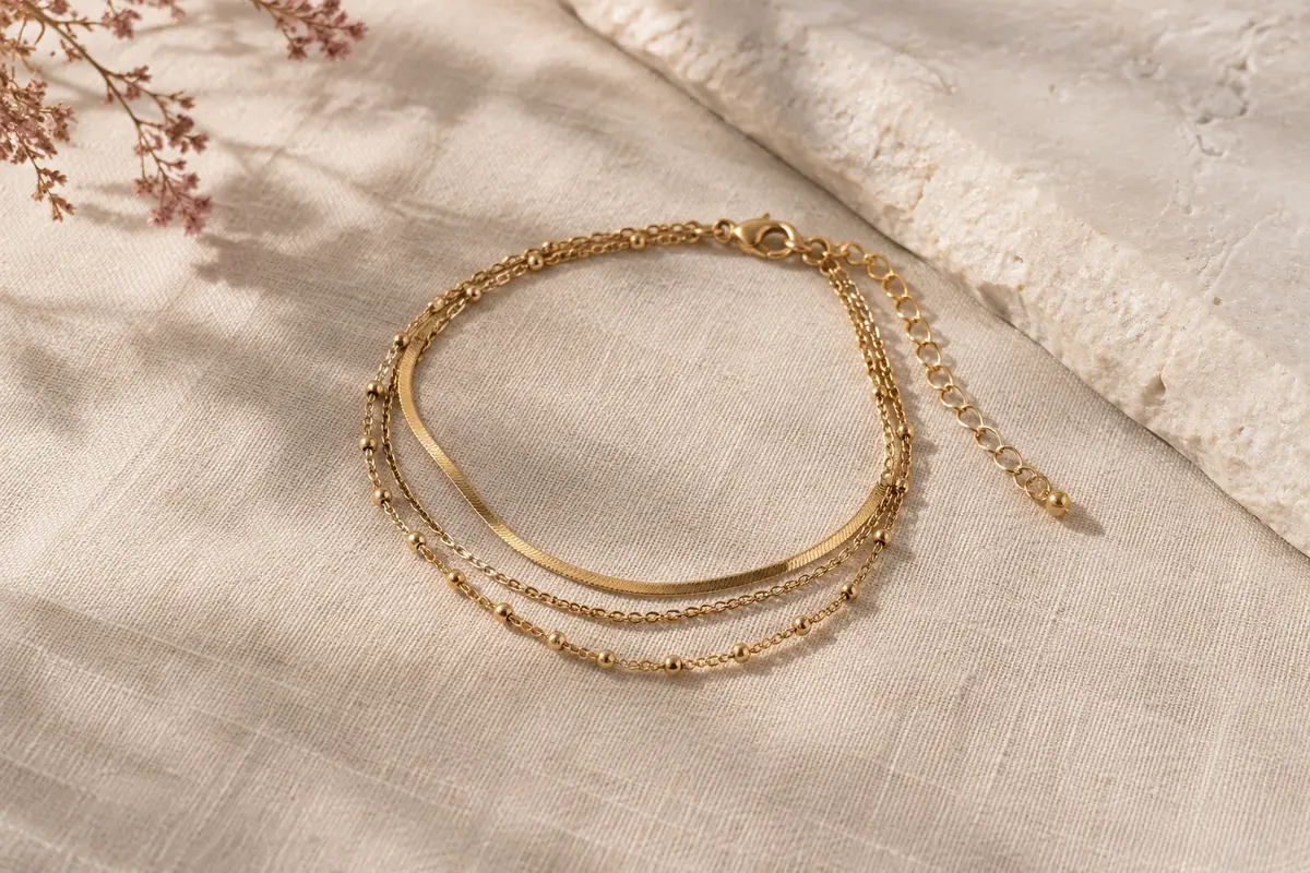 Bracelet de cheville Kenivira doré à plusieurs couches, posé sur du lin naturel clair.