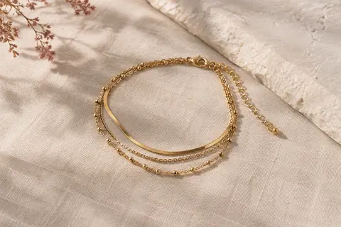 Bracelet de cheville Kenivira doré à plusieurs couches, posé sur du lin naturel clair.