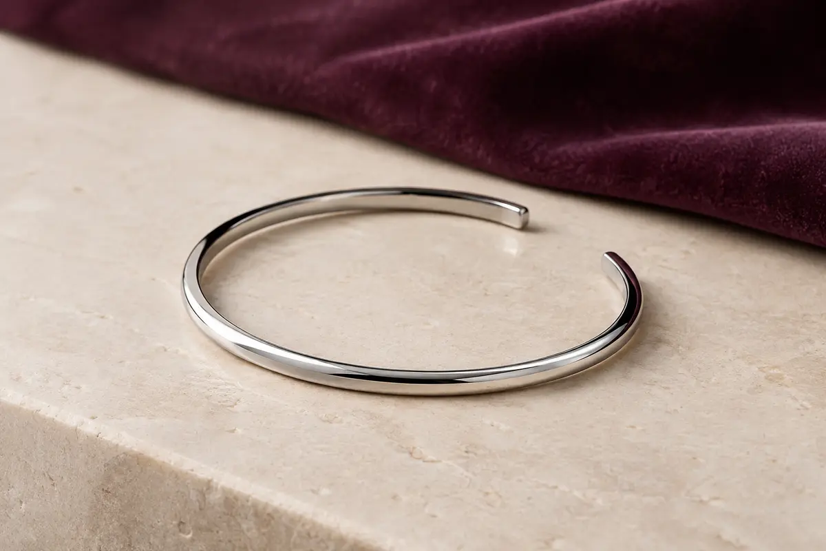 Bracelet jonc ouvert en acier inoxydable poli, posé sur une surface minérale ivoire.