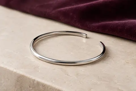 Bracelet jonc ouvert en acier inoxydable poli, posé sur une surface minérale ivoire.