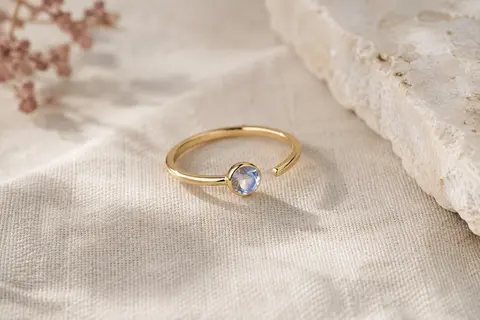 Bague ouverte dorée à pierre claire aux reflets bleutés, présentée sur du lin naturel.