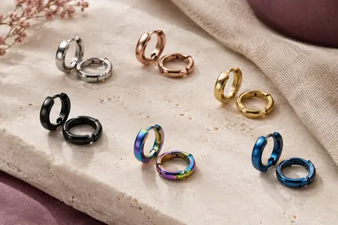 Petites créoles en acier aux finitions argentée, or rose et colorées, disposées sur une surface claire et du lin.