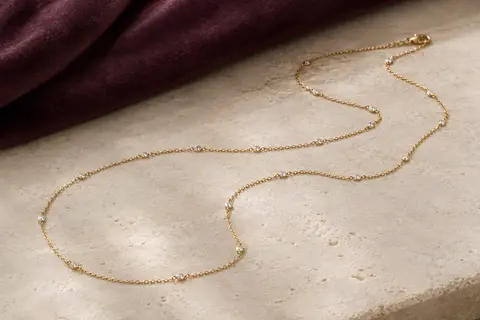 Sautoir doré en laiton plaqué or, ponctué de zircons et disposé sur une surface claire avec du velours prune.
