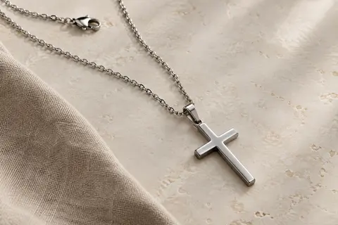 Collier en acier inoxydable à chaîne rolo et pendentif croix argentée posé sur une surface claire.