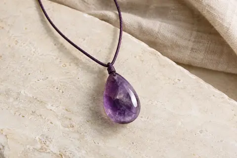 Pendentif en améthyste taillé en goutte sur un long cordon ciré, posé sur une surface minérale claire.