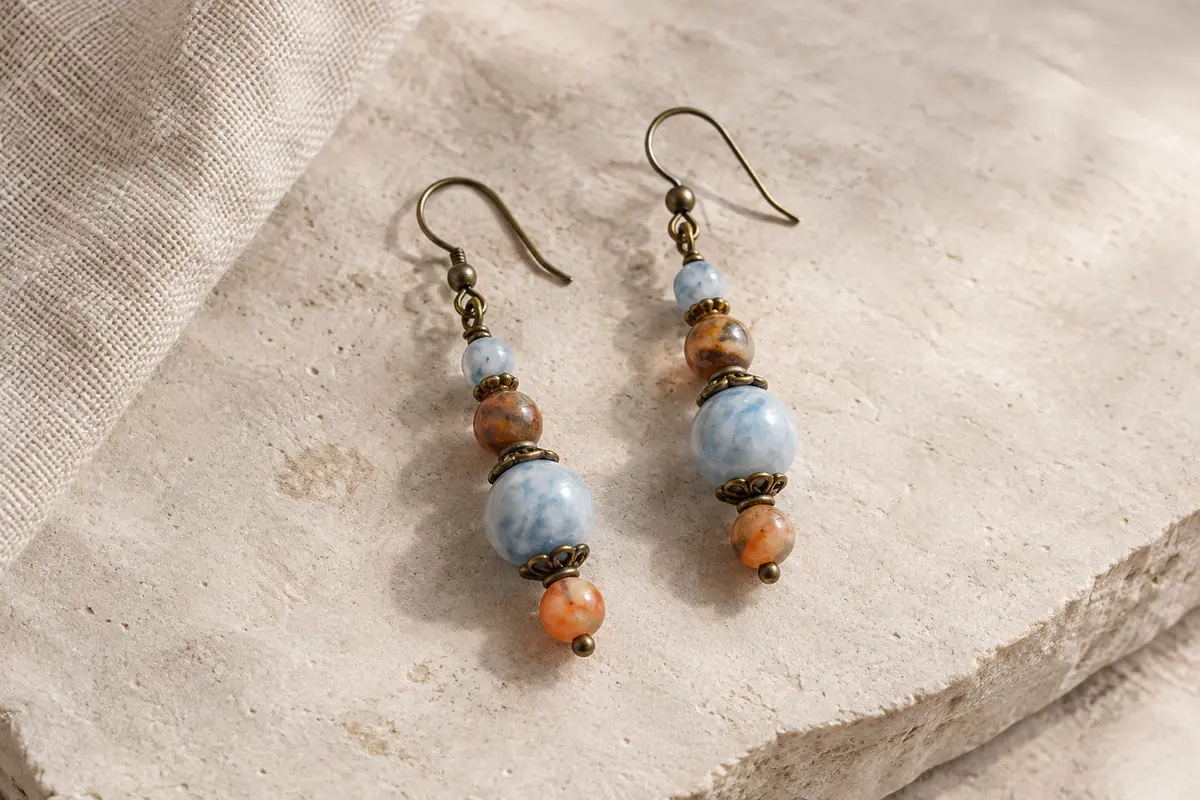 Boucles pendantes en perles de jade teinté bleu, marron et orange, posées sur une surface minérale claire.