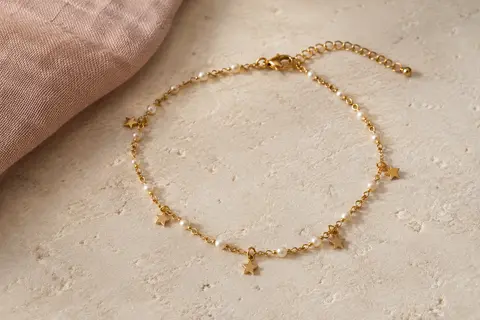 Bracelet de cheville doré à perles et étoiles, posé seul sur une surface minérale claire.