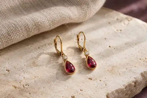 Boucles pendantes dorées à goutte rouge et petite pierre ronde, posées sur une surface minérale claire